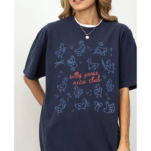 May include: Navy blue t-shirt with a whimsical design featuring cartoon ducks and the text "silly goose nicu club" in pink and blue. The shirt has a relaxed fit and short sleeves.