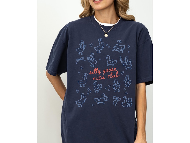 May include: Navy blue t-shirt with a whimsical design featuring cartoon ducks and the text "silly goose nicu club" in pink and blue. The shirt has a relaxed fit and short sleeves.