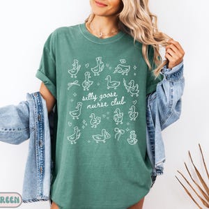 May include: Light green t-shirt with the text "Silly Goose Nurse Club" and white illustrations of geese wearing nurse hats and holding medical items. The shirt is a relaxed fit.