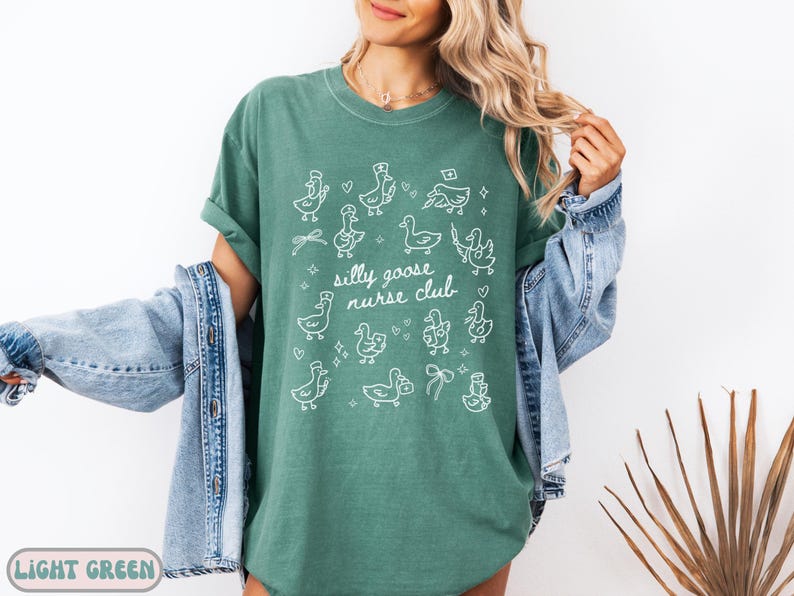 May include: Light green t-shirt with the text "Silly Goose Nurse Club" and white illustrations of geese wearing nurse hats and holding medical items. The shirt is a relaxed fit.