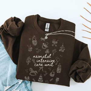 K&ouml;nnte beinhalten: Ein braunes Sweatshirt mit dem Text "neonatal intensive care unit" und verschiedenen Baby-Illustrationen in Wei&szlig;.