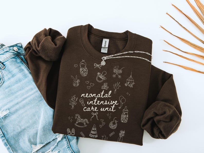 K&ouml;nnte beinhalten: Ein braunes Sweatshirt mit dem Text "neonatal intensive care unit" und verschiedenen Baby-Illustrationen in Wei&szlig;.
