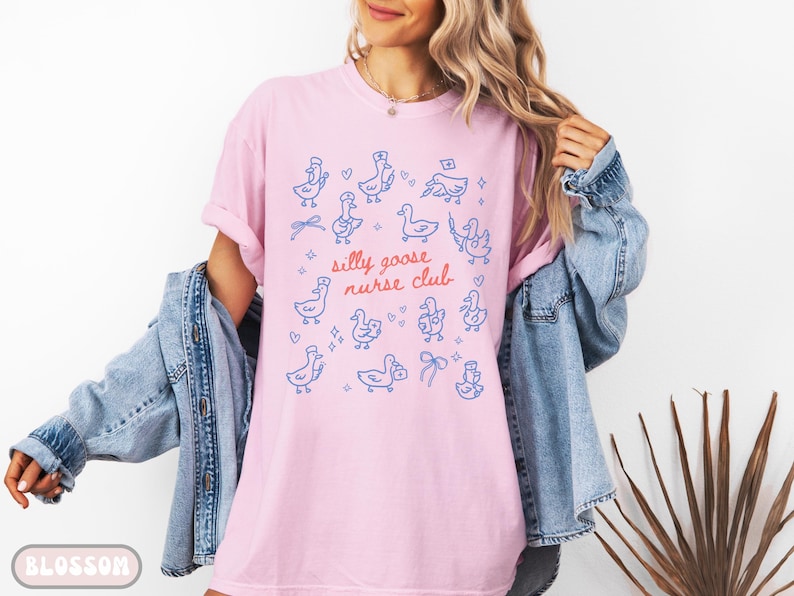 May include: Pink t-shirt with a blue illustration of geese and the text "silly goose nurse club." The design includes geese wearing nurse hats and holding medical symbols. The shirt is a soft, comfortable material.