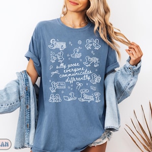 May include: Blue jean colored t-shirt with white graphic illustrations of ducks and the phrase "silly goose everyone communicates differently." The shirt has rolled-up sleeves and a relaxed fit. The words "BLUE JEAN" are at the bottom left.
