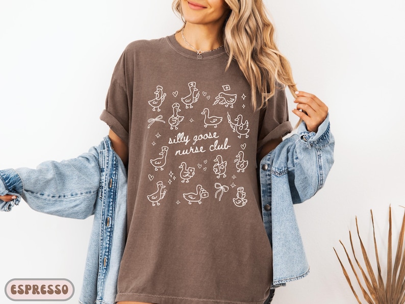 May include: Espresso-colored t-shirt with the text "Silly Goose Nurse Club" and white cartoon goose illustrations. The shirt is a relaxed fit and has rolled-up sleeves. The model is wearing a denim jacket.