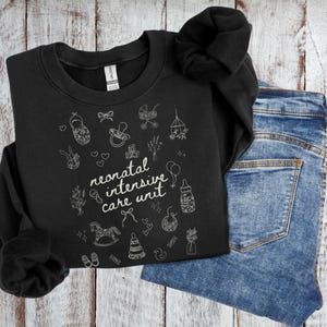 K&ouml;nnte beinhalten: Schwarzer Pullover mit wei&szlig;em Text "neonatal intensive care unit" und wei&szlig;en Illustrationen von Babyartikeln wie einem Schaukelpferd, einer Rassel und einer Babyflasche.