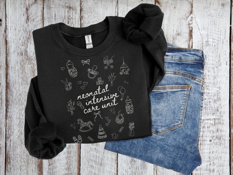 K&ouml;nnte beinhalten: Schwarzer Pullover mit wei&szlig;em Text "neonatal intensive care unit" und wei&szlig;en Illustrationen von Babyartikeln wie einem Schaukelpferd, einer Rassel und einer Babyflasche.