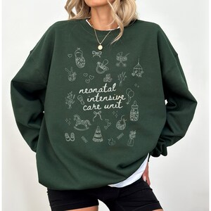 K&ouml;nnte beinhalten: Ein dunkelgr&uuml;ner Pullover mit einem wei&szlig;en Design aus babybezogenen Symbolen und dem Text "neonatal intensive care unit".