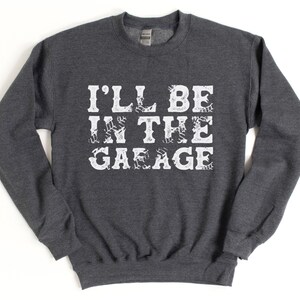 May include: A dark gray sweatshirt with the text "I'LL BE IN THE GARAGE" printed in white. The text is stylized with a tire tread pattern.