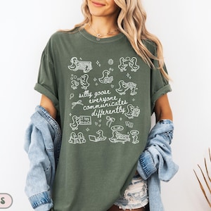 May include: Moss green t-shirt with white graphic illustrations of ducks and the text "silly goose everyone communicates differently." The shirt has short sleeves and a relaxed fit. The shirt is made of a soft, comfortable material.