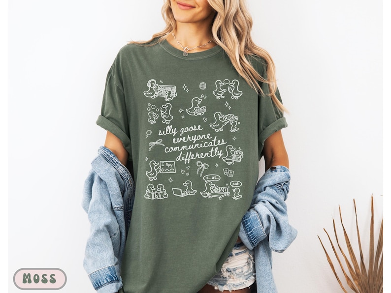 May include: Moss green t-shirt with white graphic illustrations of ducks and the text "silly goose everyone communicates differently." The shirt has short sleeves and a relaxed fit. The shirt is made of a soft, comfortable material.