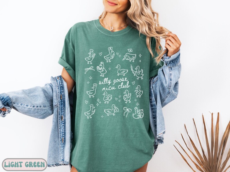 May include: Light green t-shirt with white illustrations of geese and the text "silly goose nicu club". The shirt has a crew neck and short sleeves. The design includes geese wearing hats and holding medical equipment.