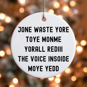 May include: A white, round Christmas ornament with a gold string for hanging. The ornament features black text that reads: "JONE WASTE YORE TOYE MONME YORALL REDIII THE VOICE INSOIDE MOYE YEDD". The background is blurred with bokeh lights.