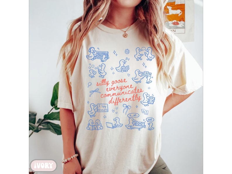 May include: Ivory-colored t-shirt with a blue graphic of ducks and the text "silly goose everyone communicates differently." The design includes ducks, bows, and speech bubbles. The shirt is a casual, comfortable style.