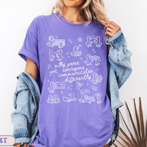 May include: Violet-colored t-shirt with white graphic illustrations of ducks and text that reads "silly goose everyone communicates differently." The shirt has short sleeves and a relaxed fit. A denim jacket is worn over the shirt.