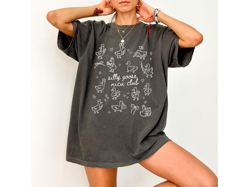 May include: Dark gray t-shirt with a whimsical design featuring cartoon geese and the text "silly goose nicu club" in white. The shirt has a relaxed fit and short sleeves, suitable for casual wear.