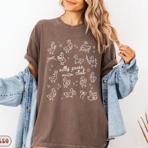 May include: Espresso-colored t-shirt with a white graphic of cartoon geese and the text "silly goose nicu club." The shirt has short sleeves and a relaxed fit. The model is wearing a denim jacket.