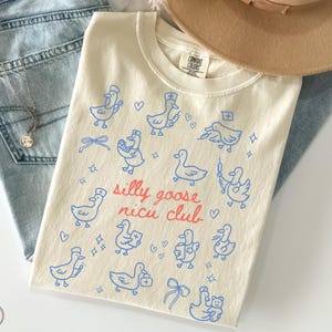 May include: Ivory-colored t-shirt with a blue cartoon goose design and the text "silly goose nicu club" in red. The shirt is folded on top of blue jeans and a tan hat.