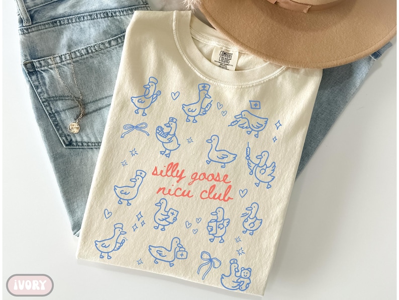 May include: Ivory-colored t-shirt with a blue cartoon goose design and the text "silly goose nicu club" in red. The shirt is folded on top of blue jeans and a tan hat.