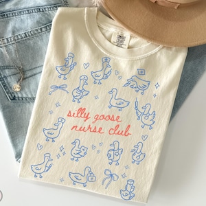 May include: Ivory-colored t-shirt with blue cartoon geese in various nurse-themed poses. The text "silly goose nurse club" is in red script. The shirt is laid on top of blue jeans and a tan hat.