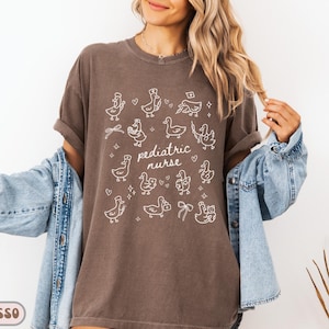 May include: Espresso-colored t-shirt with white illustrations of ducks and the words "pediatric nurse." The ducks are depicted in various poses, some wearing nurse hats. The shirt is paired with a denim jacket.