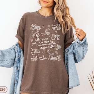 May include: Espresso-colored t-shirt with a white graphic of cartoon geese and the text "silly goose everyone communicates differently." The design includes speech bubbles, bows, and other decorative elements. The shirt has short sleeves and a relaxed fit.