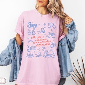 May include: Pink t-shirt with a blue illustration of ducks and the text "silly goose everyone communicates differently." The shirt has a relaxed fit and is paired with a denim jacket. The word "BLOSSOM" is printed at the bottom.