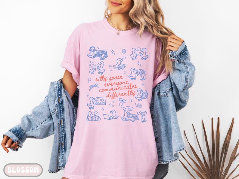 May include: Pink t-shirt with a blue illustration of ducks and the text "silly goose everyone communicates differently." The shirt has a relaxed fit and is paired with a denim jacket. The word "BLOSSOM" is printed at the bottom.