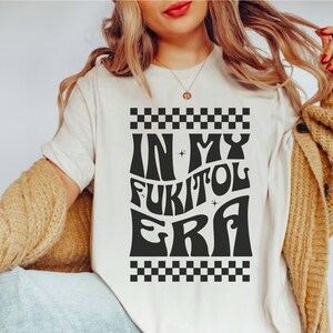 May include: Cream-colored t-shirt with the text "IN MY FUKITOL ERA" in a black, retro font. The design includes a checkered pattern at the top and bottom. The shirt is a casual, everyday wear item.