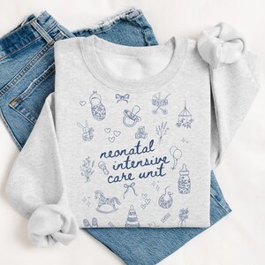 K&ouml;nnte beinhalten: Ein graues Sweatshirt mit einem blauen Design aus babybezogenen Symbolen und dem Text "neonatal intensive care unit".
