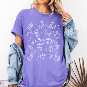 May include: A violet-colored t-shirt with a white graphic of cartoon geese and the text "silly goose nicu club." The shirt has short sleeves and a relaxed fit. The design includes various goose illustrations with hearts and bows.