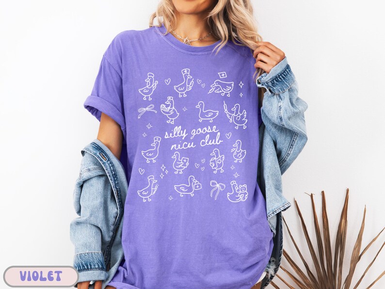 May include: A violet-colored t-shirt with a white graphic of cartoon geese and the text "silly goose nicu club." The shirt has short sleeves and a relaxed fit. The design includes various goose illustrations with hearts and bows.