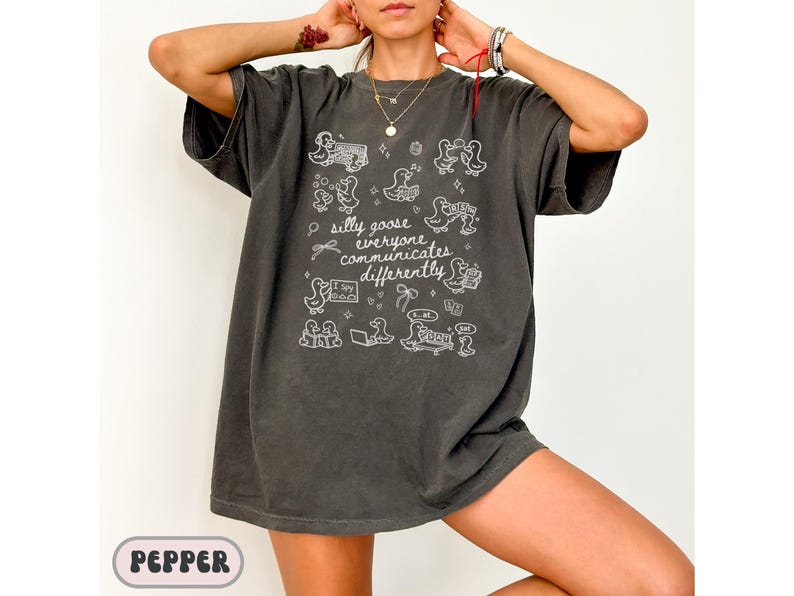 May include: Dark gray t-shirt with white graphic illustrations of ducks and the text "silly goose everyone communicates differently." The shirt has a relaxed fit and short sleeves. The shirt is a casual wear item.