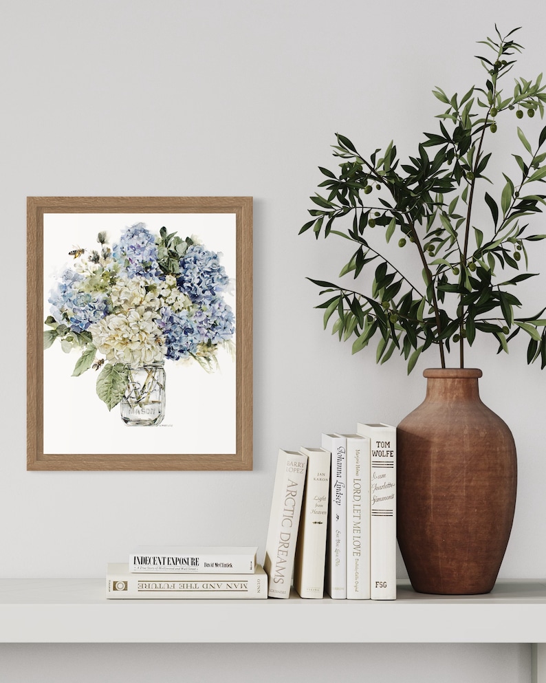 Pode incluir: Uma pintura em aquarela emoldurada de um buqu&ecirc; de hort&ecirc;nsias azuis e brancas em um frasco de vidro transparente. As flores est&atilde;o dispostas em um vaso com uma moldura de madeira marrom. A pintura est&aacute; em uma parede branca com uma oliveira em vaso e uma prateleira com livros.