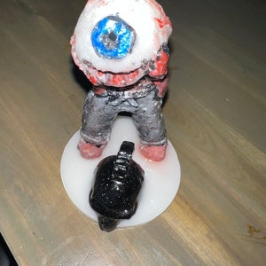 May include: A handmade figurine of a creature with a large, white eyeball and red skin. The creature is wearing grey pants and is standing on a white base. A black skull with glitter is at the base of the figurine.