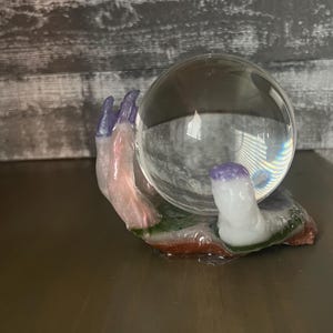 May include: A glass orb sits in the palm of a hand sculpted from resin. The hand is painted in shades of pink, purple, and green. The orb is clear and reflects the surrounding environment.