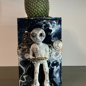 May include: A gray alien figurine holding a tray of gears, set against a black and white marbled background with a green cactus in a pot on top.