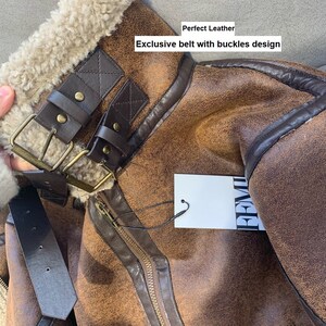 Op de afbeelding: Close-up van een bruin leren jack met een bontkraag en een bruine leren riem met gespen. Het jack heeft een ritssluiting en een label met de tekst "PREMIUM". De tekst "Perfect Leather" en "Exclusive belt with buckles design" is zichtbaar op de afbeelding.
