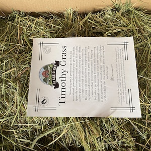 Op de afbeelding: Een close-up van een doos gevuld met groen Timothy-gras hooi. Een wit papier met de tekst "FLAKE O HAY Timothy Grass" is zichtbaar bovenop het hooi.