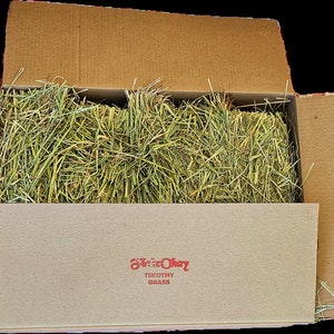 Op de afbeelding: Een kartonnen doos gevuld met groen Timothy-gras hooi. De doos is open en het hooi is zichtbaar van binnen. De doos heeft een rood label met de tekst "Sunshine Hay" en "Timothy Grass".