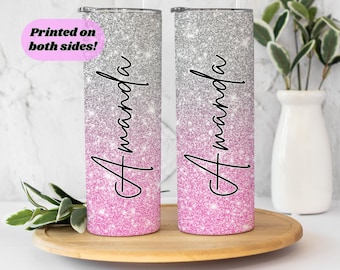 May include: Two silver and pink glitter tumblers with the name "Amanda" printed on each. The tumblers are on a wooden surface.