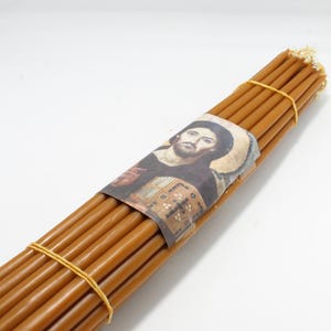 Puede incluir: Un paquete de 25 velas de cera de abeja marrón atadas con gomas elásticas. Las velas están envueltas en una etiqueta de papel con un icono religioso de Jesucristo.