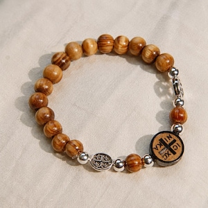 May include: A stretch bracelet featuring round brown wood beads with visible grain patterns, interspersed with small silver-toned metal beads. It includes a circular dark wood charm engraved with a cross and the text IC XC NIKA, plus a small oval silver charm.