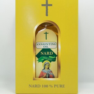 May include: A yellow box with a cross-shaped aperture, displaying a bottle of "Anointing Oil Nard 100% Pure." The bottle label has a green banner and an illustration of a woman. The text "NARD 100% PURE" is printed on the box.