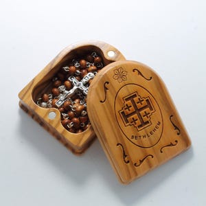 Pode incluir: Uma caixa de madeira com tampa articulada, contendo um rosário de madeira castanho com uma cruz de prata. A caixa é gravada com as palavras "Bethlehem" e uma cruz.