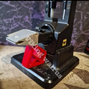 Może przedstawiać: Czarny ostrzał do noży z srebrnym ostrzem i czerwoną plastikową podstawą. Ostrzał ma dźwignię i pokrętło do regulacji kąta ostrza. Ostrzał jest oznaczony "SHARP" na podstawie.