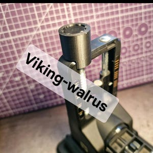 Może przedstawiać: Czarne metalowe narzędzie z cylindryczną górą i gwintowanym prętem. Na obrazie widnieje tekst "Viking-walrus". Narzędzie jest umieszczone na fioletowym tle w kratkę.