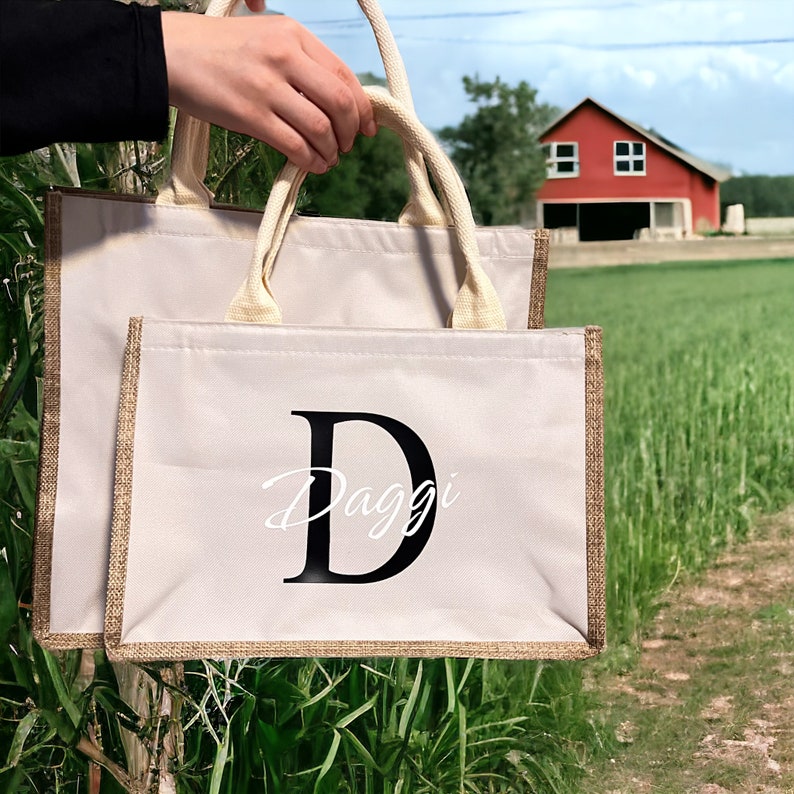 K&ouml;nnte beinhalten: Zwei beigefarbene Tragetaschen mit Jutebesatz und Seilgriffen werden hochgehalten. Die Taschen tragen den Namen "Daggi" in einem schwarzen Gro&szlig;buchstaben "D" und in Schreibschrift. Im Hintergrund eine rote Scheune und ein gr&uuml;nes Feld.