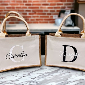K&ouml;nnte beinhalten: Zwei beigefarbene Tragetaschen mit braunen Verzierungen und cremefarbenen Griffen. Eine Tasche hat den Namen "Carolin" in einer Schreibschrift mit einem gro&szlig;en "C" aufgedruckt. Die andere Tasche hat den Namen "Daggi" in einer Schreibschrift mit einem gro&szlig;en "D" aufgedruckt.
