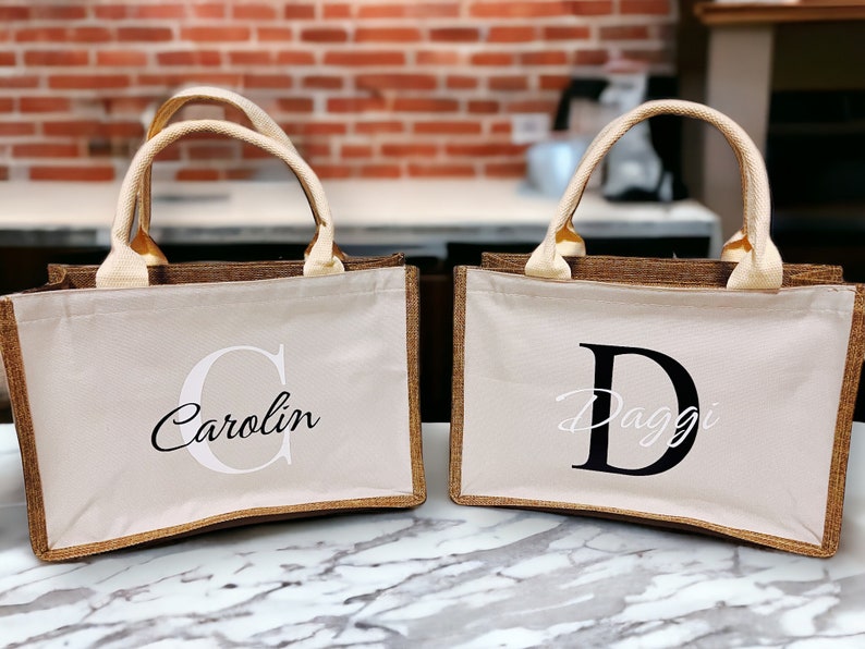 K&ouml;nnte beinhalten: Zwei beigefarbene Tragetaschen mit braunen Verzierungen und cremefarbenen Griffen. Eine Tasche hat den Namen "Carolin" in einer Schreibschrift mit einem gro&szlig;en "C" aufgedruckt. Die andere Tasche hat den Namen "Daggi" in einer Schreibschrift mit einem gro&szlig;en "D" aufgedruckt.